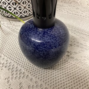 May include: A blue speckled fragrance lamp with a black top. The lamp has a rounded, bulbous shape and sits on a white lace tablecloth. The top is a vented, cylindrical shape.