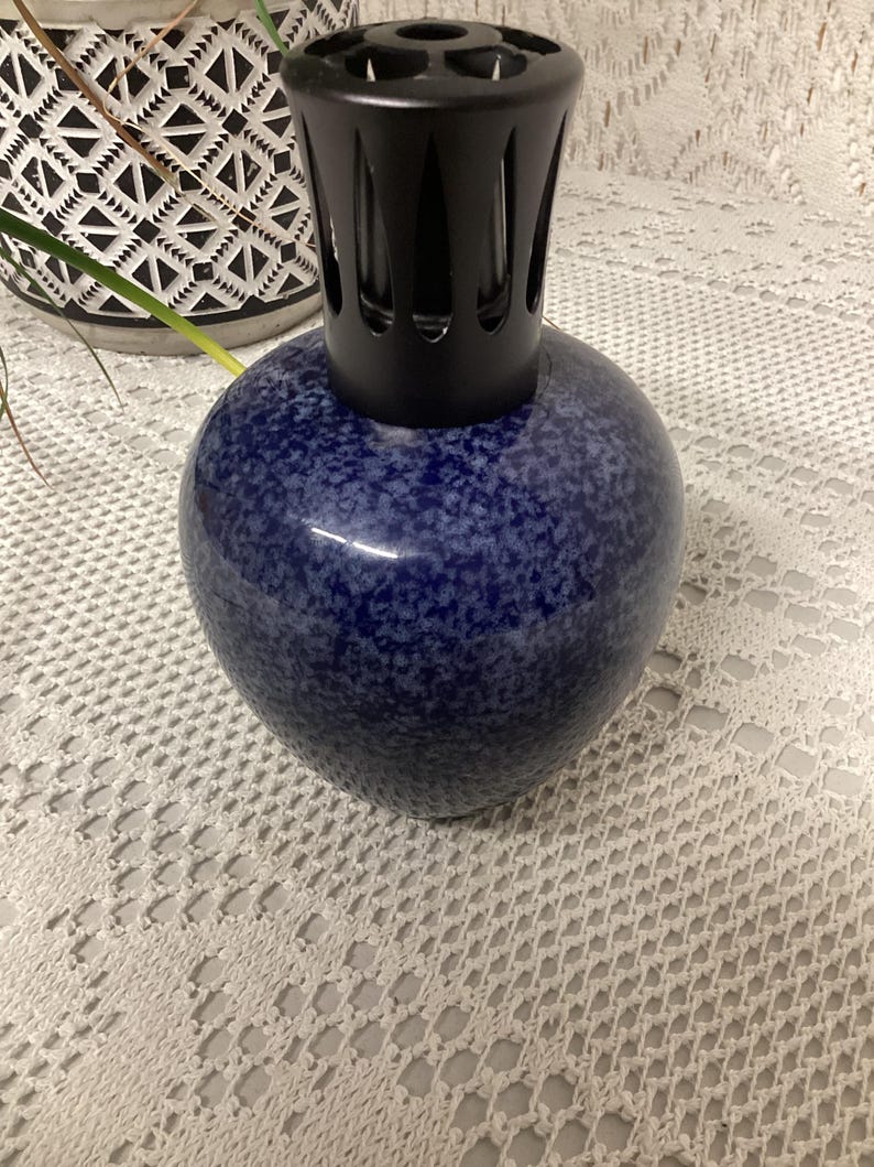 May include: A blue speckled fragrance lamp with a black top. The lamp has a rounded, bulbous shape and sits on a white lace tablecloth. The top is a vented, cylindrical shape.