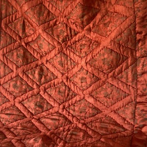 May include: A quilted textile in a warm orange hue, featuring a diamond pattern with floral accents. The fabric has a subtle sheen, and the edges are finished with a satin border. This textile could be a throw, blanket, or decorative piece.