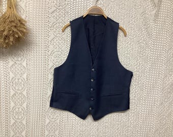 May include: A navy blue waistcoat with a V-neck and button closure. It features two front pockets and is suspended on a wooden hanger. The backdrop is a white, patterned textile.