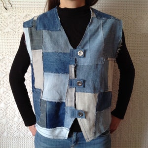May include: A patchwork denim vest in various shades of blue. The vest features a V-neck and is fastened with three silver buttons. The design incorporates rectangular and square denim patches, creating a unique, textured look. The vest is sleeveless.