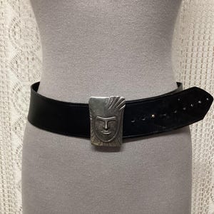 May include: A black leather belt with a silver-colored buckle featuring a stylized face design. The belt is wide and has several holes for adjustment. The buckle is rectangular and has a detailed face.