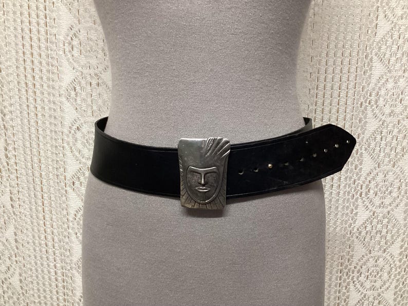 May include: A black leather belt with a silver-colored buckle featuring a stylized face design. The belt is wide and has several holes for adjustment. The buckle is rectangular and has a detailed face.