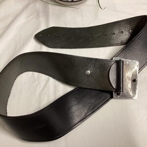 May include: A black leather belt with a silver-colored rectangular buckle. The belt is wide and has several holes for adjustment. The buckle is a simple, rectangular design.