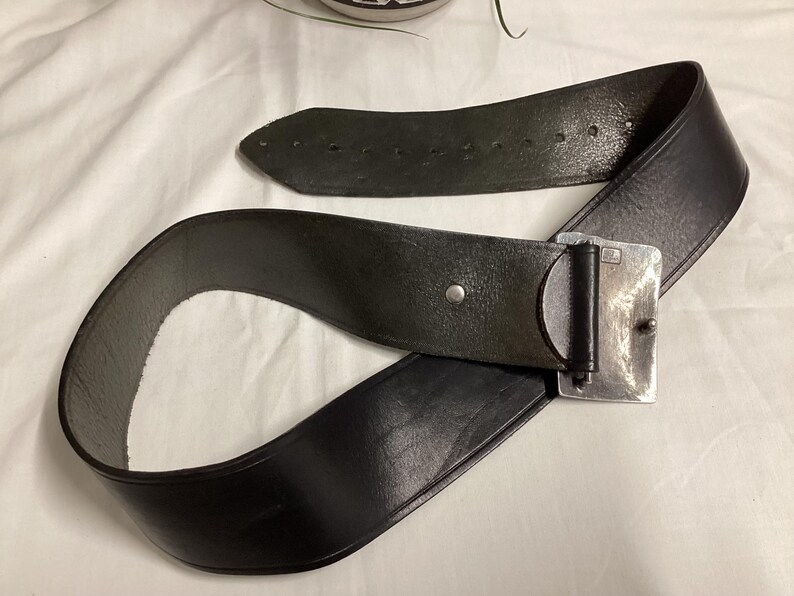 May include: A black leather belt with a silver-colored rectangular buckle. The belt is wide and has several holes for adjustment. The buckle is a simple, rectangular design.