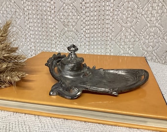 May include: Antique silver-toned inkwell with a decorative design, featuring a lidded ink pot and an attached tray. The inkwell is placed on a vintage book with a wheat stalk in the background. The inkwell's ornate details and aged appearance suggest a vintage aesthetic.