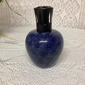 May include: A blue speckled ceramic oil diffuser with a black top. The diffuser has a rounded body and a textured surface. The top is a black, open-slatted design. The diffuser is sitting on a white lace tablecloth.