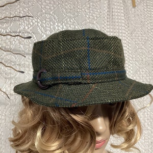 Può includere: Un cappello a secchiello in tweed verde, marrone e blu a quadri con una fibbia in pelle marrone.
