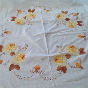 Peut inclure: Nappe blanche avec un motif circulaire brodé de fleurs jaunes et brunes. Les fleurs sont disposées en un motif répétitif autour du bord de la nappe.