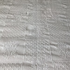 May include: White quilted fabric with a repeating pattern of squares. Each square features a different textured design, including floral and dotted patterns. The overall effect is a classic, textured look.