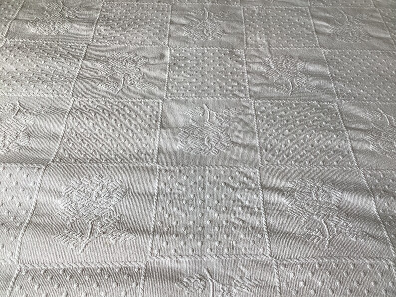 May include: White quilted fabric with a repeating pattern of squares. Each square features a different textured design, including floral and dotted patterns. The overall effect is a classic, textured look.