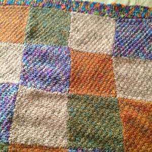Könnte beinhalten: Eine handgestrickte Decke mit quadratischem Muster. Die Decke zeigt ein Patchwork aus Quadraten in verschiedenen Farben, darunter Orange, Grün, Lila und Creme. Die Ränder sind mit einem farbenfrohen, strukturierten Rand versehen.
