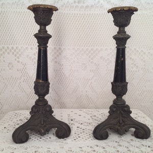 May include: A pair of antique candle holders with intricate designs. The candle holders feature a dark central column, possibly black, with decorative bases and tops. The tops have a gold-coloured rim.
