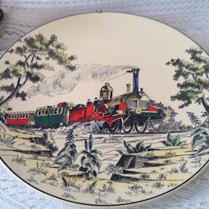 Puede incluir: Un plato de cerámica vintage con una ilustración colorida de un tren de vapor que viaja a través de un bosque verde. El tren es rojo, verde y negro con humo blanco saliendo de la chimenea. El plato tiene un fondo crema con un borde marrón.