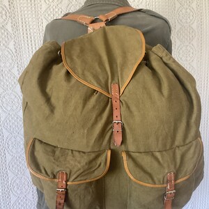 May include: A large, green canvas backpack with brown leather straps and buckles. The backpack has two large pockets on the front.