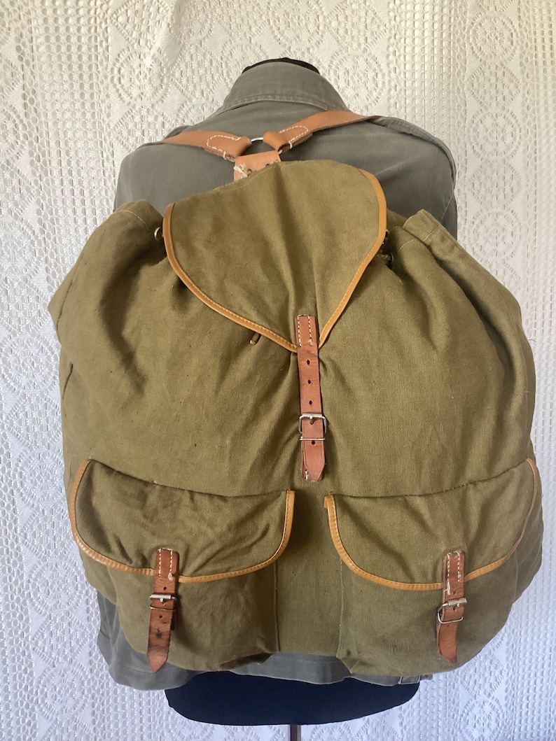 May include: A large, green canvas backpack with brown leather straps and buckles. The backpack has two large pockets on the front.