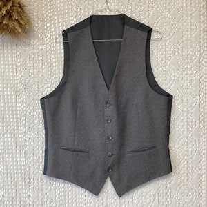 Peut inclure: Un gilet gris avec une doublure noire et cinq boutons. Le gilet a deux poches sur le devant.