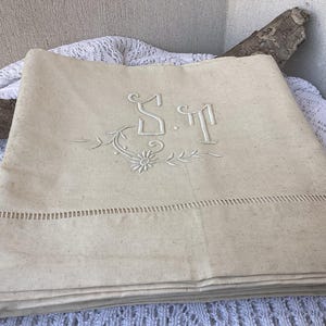 Può includere: Lenzuolo in lino beige con iniziali ricamate "S.M" e dettagli floreali. Il lenzuolo presenta un bordo traforato decorativo sul bordo inferiore. Il tessuto è piegato, mostrando la sua trama e il ricamo dettagliato.