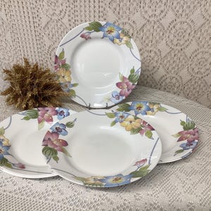 Könnte beinhalten: Set aus vier weißen Keramik-Dinnertellern mit einem Blumendesign mit blauen, rosa und gelben Blumen. Die Teller haben einen erhöhten Rand und einen leicht gewellten Rand.