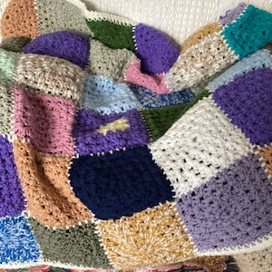 Peut inclure: Une couverture en patchwork crochetée avec des carrés de différentes couleurs, notamment violet, rose, bleu, vert et marron.