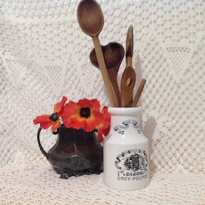 Könnte beinhalten: Eine weiße Keramikdose mit einem schwarz-weißen Etikett mit der Aufschrift "Dijon Grey-Poupon" ist mit Holzlöffeln gefüllt. Die Dose steht auf einer weißen Spitzentischdecke mit einer kleinen, dunklen Metallvase mit orangefarbenen Blumen.