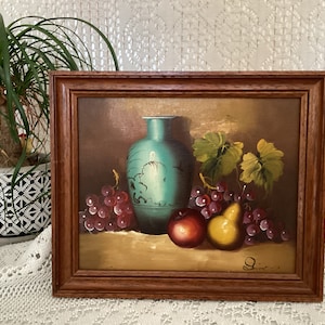 Könnte beinhalten: Ein gerahmtes Stillleben mit einer blauen und weißen Vase mit Trauben, einer Birne und einem Apfel. Das Gemälde ist vor einem braunen Hintergrund und hat einen goldfarbenen Rahmen.