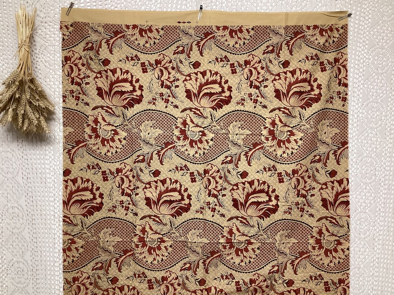 May include: A beige fabric with a repeating pattern of red stylized flowers and leaves. The fabric has a woven texture.