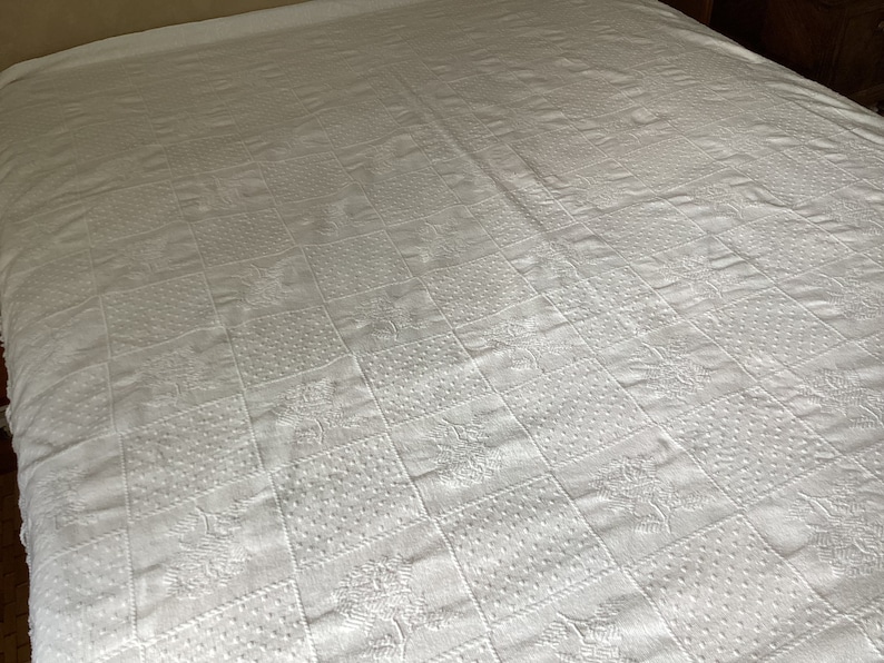 May include: White bedspread with a quilted pattern. The pattern is composed of squares with various textures, including dots and floral designs. The bedspread covers the entire bed, creating a clean and classic look.