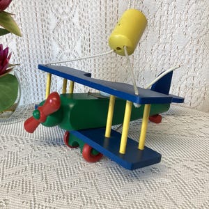 May include: A colourful wooden biplane toy, painted in blue, green, red, and yellow. The biplane features a propeller, wheels, and a yellow pilot figure attached by strings. The toy is set against a white lace background.