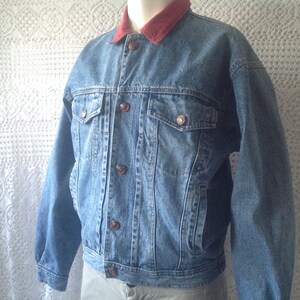 Peut inclure: Une veste en jean bleu avec un col en velours côtelé rouge. La veste a une fermeture à boutons et deux poches sur le devant.