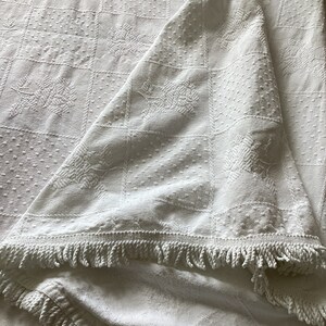 May include: White, textured blanket with a square pattern and floral designs. The blanket has a fringe trim along the edge. The fabric is folded over, showing the underside of the blanket.