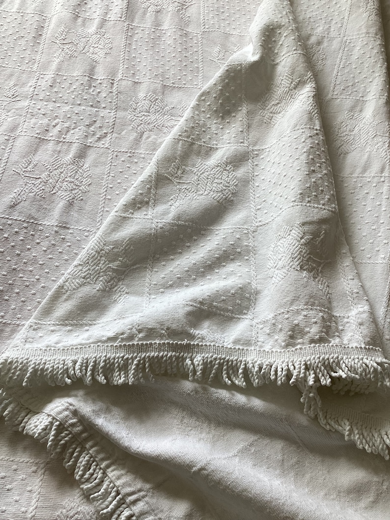 May include: White, textured blanket with a square pattern and floral designs. The blanket has a fringe trim along the edge. The fabric is folded over, showing the underside of the blanket.