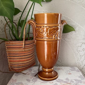 May include: A tall, brown ceramic vase with a pedestal base and two handles. The vase features a decorative band with a relief pattern of leaves and acorns. The vase is approximately 12 inches tall.