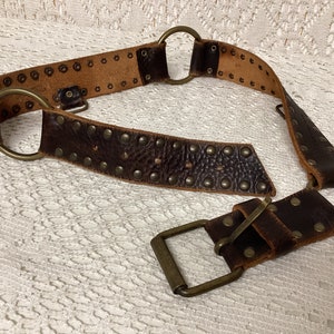 Peut inclure: Une ceinture en cuir marron avec des clous métalliques et une boucle en laiton. La ceinture a deux anneaux en D à l'extrémité.