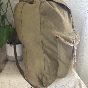 May include: A large, green canvas backpack with brown leather straps and buckles. The backpack has a flap closure and a large pocket on the front.