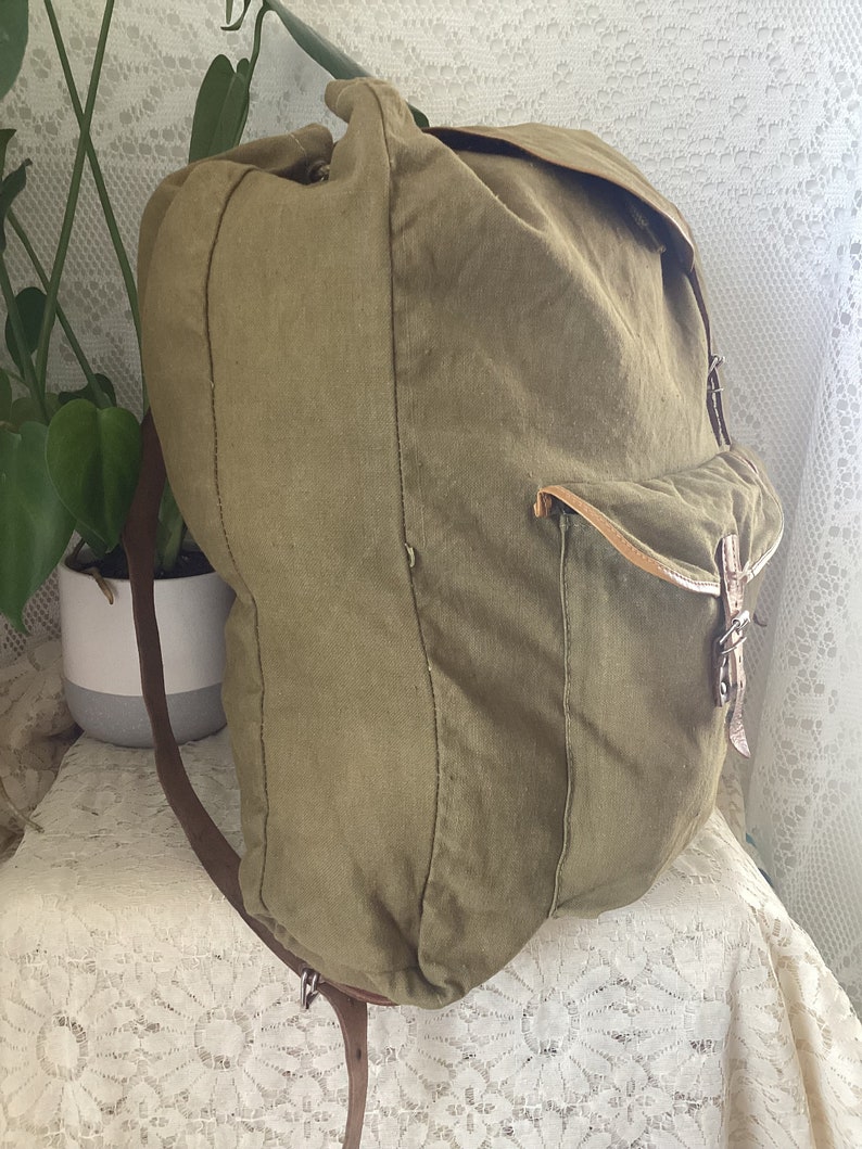 May include: A large, green canvas backpack with brown leather straps and buckles. The backpack has a flap closure and a large pocket on the front.