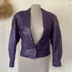 Può includere: Giacca vintage in pelle viola con chiusura a un bottone. La giacca presenta un colletto a revers, maniche lunghe e toppe decorative sulle spalle. La giacca è esposta su un manichino.