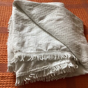 May include: A folded white blanket with a textured pattern and fringe trim. The blanket features a repeating design of squares, dots, and floral motifs. The blanket is folded on an orange textured surface.