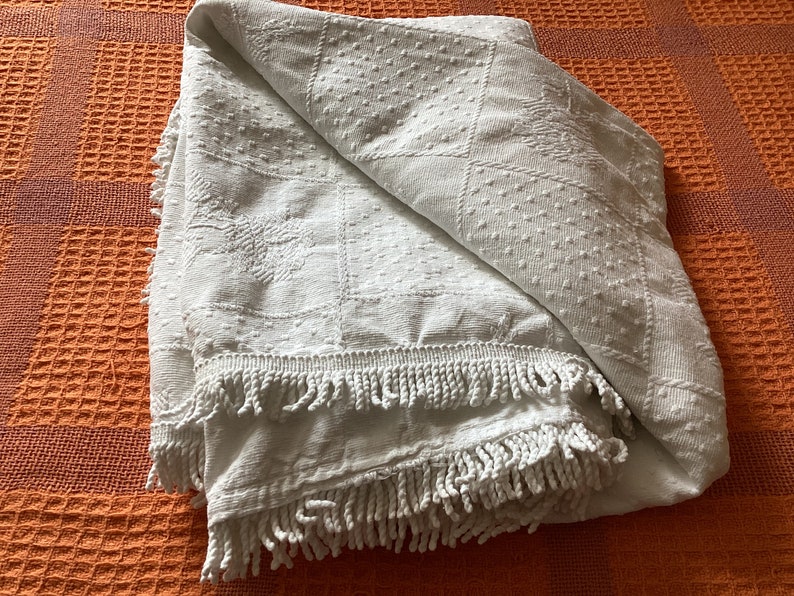May include: A folded white blanket with a textured pattern and fringe trim. The blanket features a repeating design of squares, dots, and floral motifs. The blanket is folded on an orange textured surface.