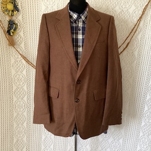 Puede incluir: Una chaqueta marrón con un cierre de botón único y dos bolsillos. La chaqueta está hecha de una tela texturizada y tiene un estilo clásico.