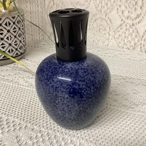 May include: A blue speckled ceramic fragrance lamp with a black top. The lamp has a rounded base and a decorative black cap with vertical slits. The lamp is sitting on a white lace tablecloth.