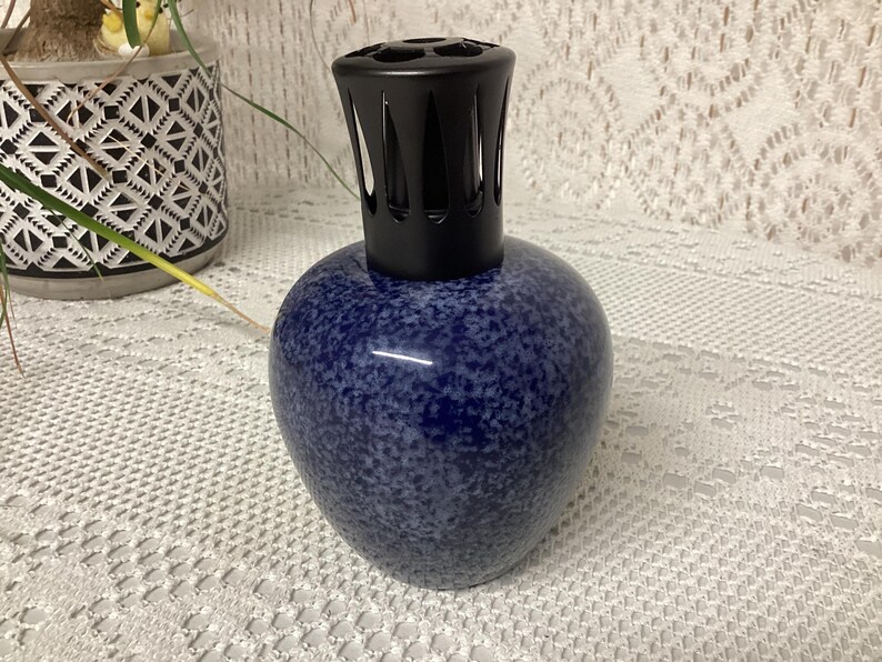 May include: A blue speckled ceramic fragrance lamp with a black top. The lamp has a rounded base and a decorative black cap with vertical slits. The lamp is sitting on a white lace tablecloth.
