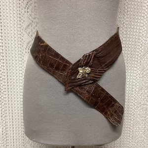 May include: A brown leather belt with a crocodile texture. The belt is wrapped around a mannequin and features a gold-coloured floral buckle with a small, clear gem in the centre. A vintage fashion accessory.