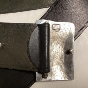 May include: Close-up of a leather belt with a silver-colored buckle. The belt is dark green and black. The buckle is rectangular with a small, stamped design and a silver-colored pin.