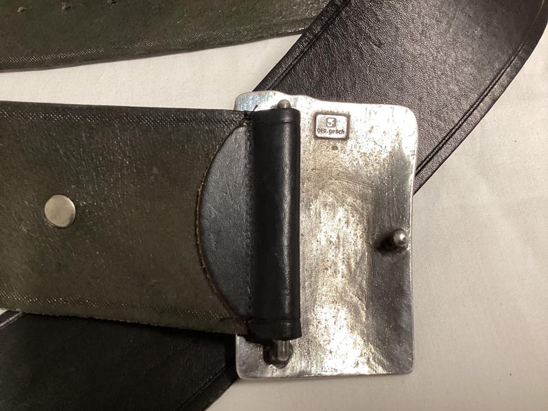 May include: Close-up of a leather belt with a silver-colored buckle. The belt is dark green and black. The buckle is rectangular with a small, stamped design and a silver-colored pin.