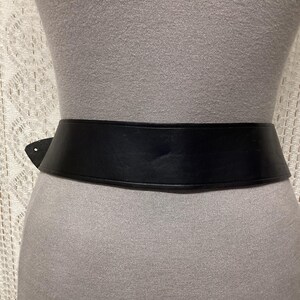 May include: A wide, black belt with a simple design. The belt is made of a smooth material and is worn around the waist. The belt is shown on a mannequin. The background is a light-colored, textured fabric.