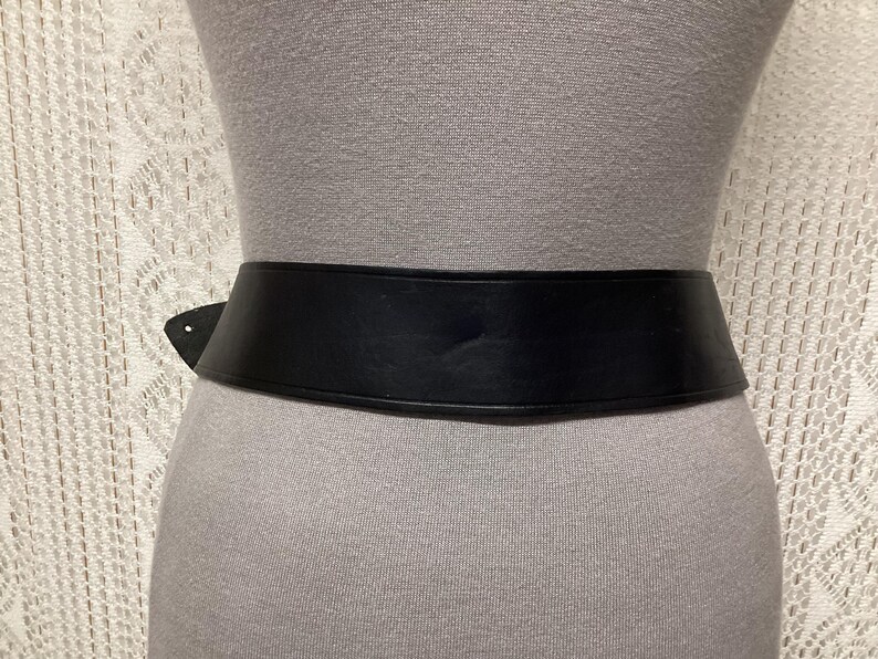 May include: A wide, black belt with a simple design. The belt is made of a smooth material and is worn around the waist. The belt is shown on a mannequin. The background is a light-colored, textured fabric.