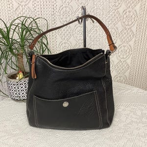 Pu&ograve; includere: Borsa a tracolla in pelle nera con chiusura superiore con cerniera e una tasca frontale. La borsa ha un manico in pelle marrone e dettagli di cuciture bianche. La borsa &egrave; su una superficie bianca.