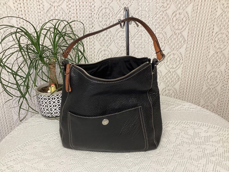 Pu&ograve; includere: Borsa a tracolla in pelle nera con chiusura superiore con cerniera e una tasca frontale. La borsa ha un manico in pelle marrone e dettagli di cuciture bianche. La borsa &egrave; su una superficie bianca.