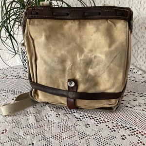Puede incluir: Un bolso de mensajero vintage de lona y cuero con un aspecto desgastado y envejecido. El bolso tiene una correa y una hebilla de cuero marrón, y una solapa con un cierre de cuero marrón.